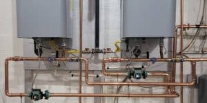 Two tankless water heaters with copper piping, gas lines, and circulation pumps installed on a wall.