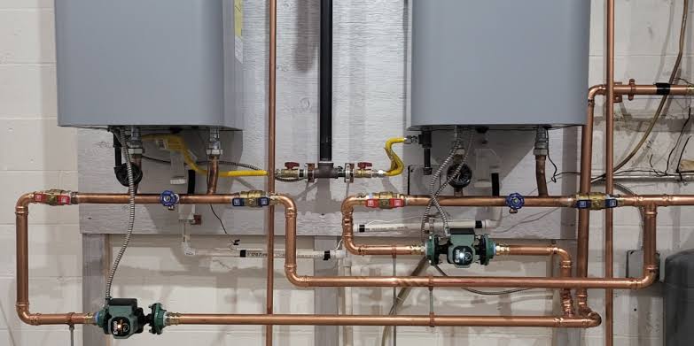 Two tankless water heaters with copper piping, gas lines, and circulation pumps installed on a wall.