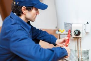 A plumber in a blue uniform uses a pipe wrench to repair a residential water heater.